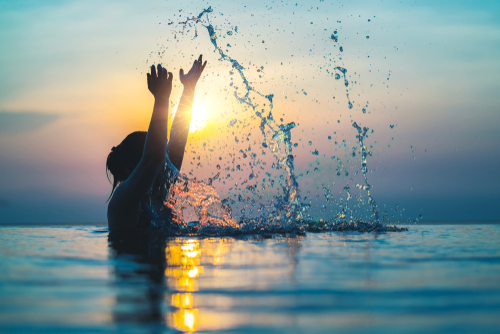 side silhouette of woman in ocean up to chest holding up arms splashing at sunset