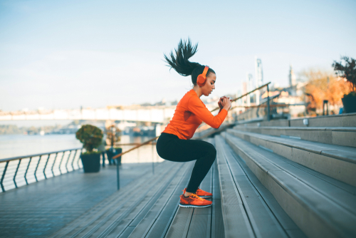woman wearing orange sweater black pants and orange sneakers doing squats on outside stairs by the ocean