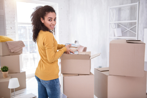 young woman smiling with afro curly hair standing by moving boxes as she moves into a white painted college dorm