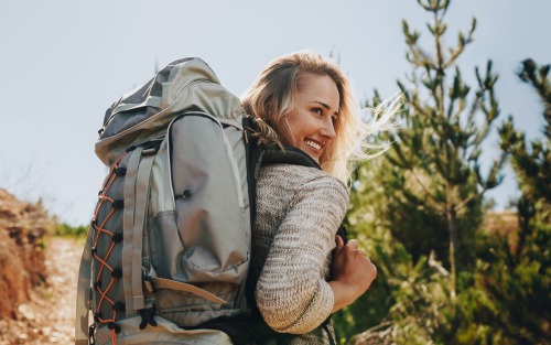 rear view shot of smiling woman looking back with backpack on her back in the outdoors surrounded by trees and nature