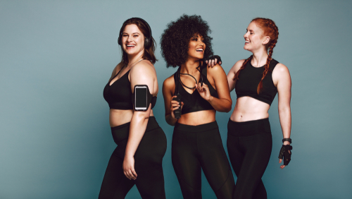 a multi ethnic group of three women dressed in black yoga pants and sports bras standing in front of a teal background smiling