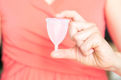a woman hand holding a clear white menstrual cup between thumb and finger in front of her the persons peach blouse in the background