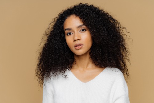 young women with natural long afro hair standing in a white v neck sweater against a plain tan colored background