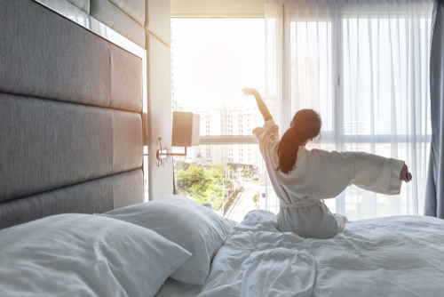 a view behind a woman sitting on the edge of a hotel bed with her arms stretched out looking out the hotel room window