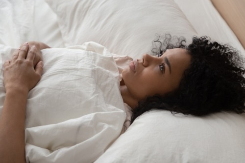 young woman with afro hair laid in a white bed staring to the ceiling looking anxious
