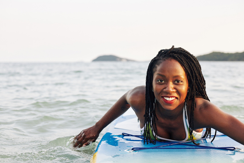 smiling african American woman with braids laid on surfboard padding towards the camera in the ocean