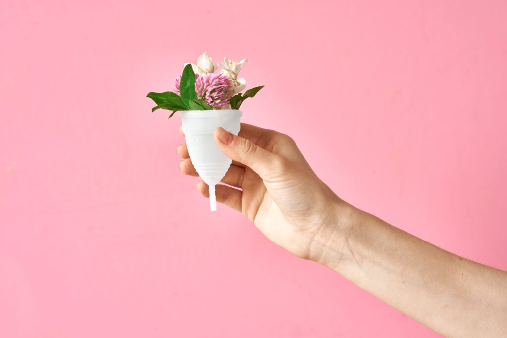 hand holding a white menstrual cup filled with flowers against a pink background