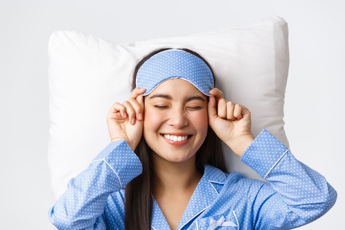 smiling and comfortable young woman laid back on a bed in blue polka dot pajamas lifting down a matching eye sleep mask