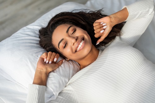 African American woman in white sweater laying back on white bed smiling with a calm and peaceful look