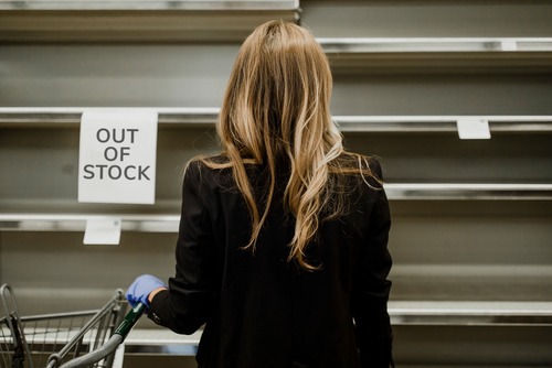 the back of a woman with long blonde hair staring at empty supermarket shelves and an out of stock sign