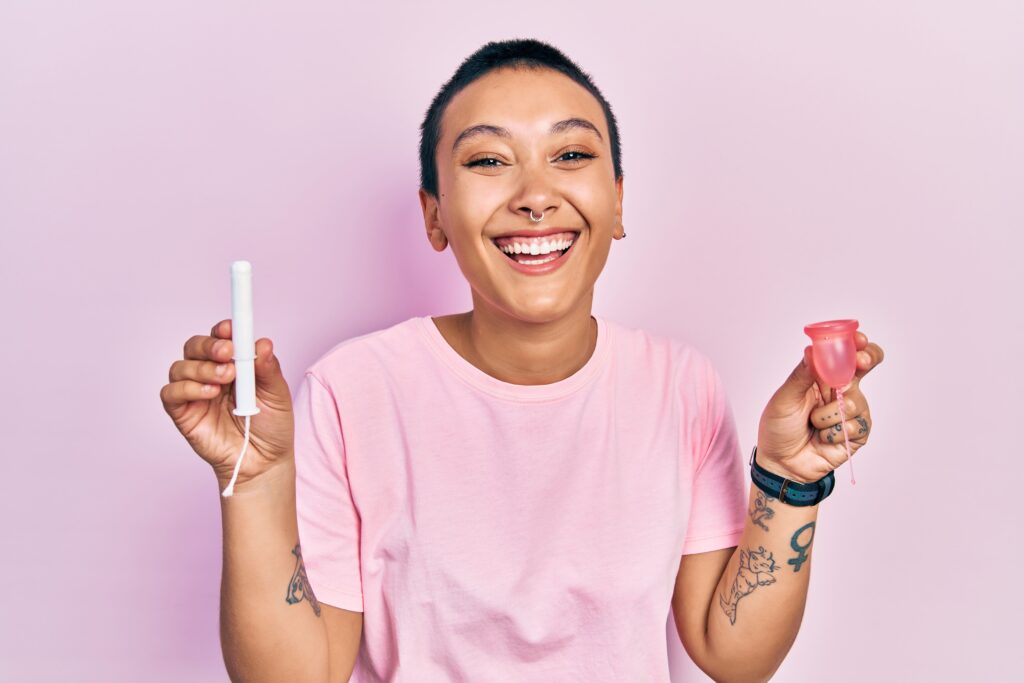 woman with short hair in pink t shirt smiling while holding up a tampon in one hand and a menstrual cup in the other hand