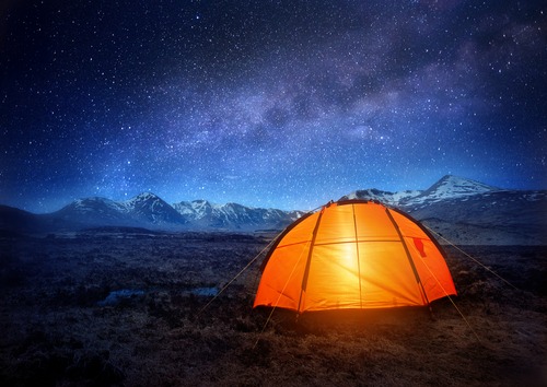 A single glowing orange tent in a barren empty place at night underneath the stars and Milky Way that create blue and purple cloud patterns in the sky