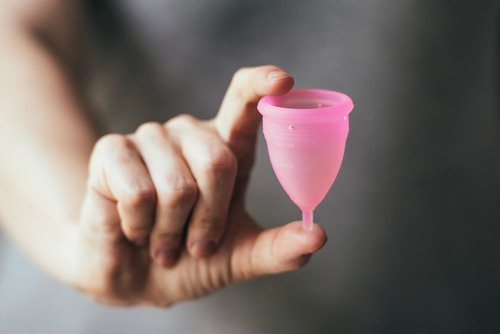 woman hand close up holding pink menstrual cup between finger and thumb against background of her gray sweater
