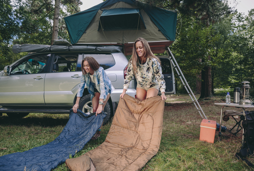 two young women in front of a tent and car laying our their sleeping bags in front of them