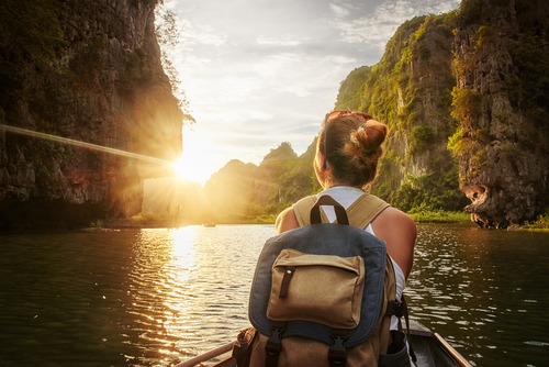 behind a young woman wearing a backpack sitting in the front of a canoe boat looking out to the view of the river and mountains and greenery at either side