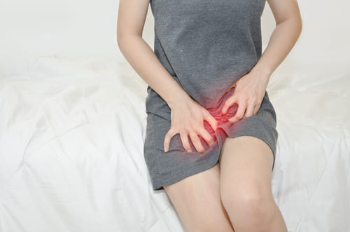 woman sitting on a bed in grey dress holding pelvic area which is glowing red indicating discomfort