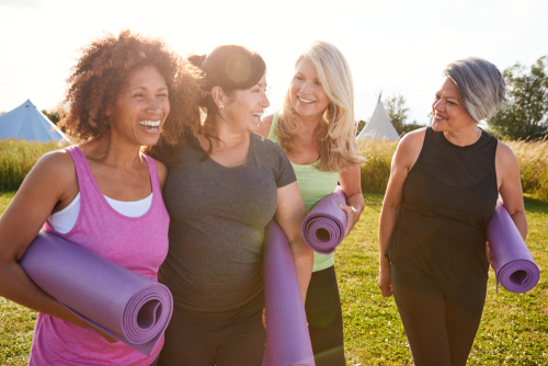 group of 4 women walking together outside in a park smiling in gym wear holding purple yoga mats