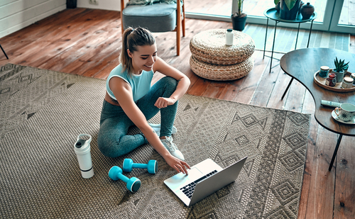 young woman sat on a rug in her living room in gym clothes with dumbbells and water next to her typing on a computer