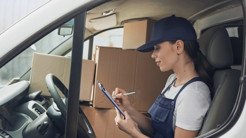 woman sitting in drivers seat of delivery van that is full of packages while looking at a clipboard