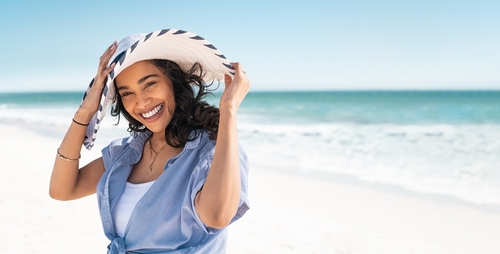 smiling woman with long dark hair wearing a white hat and a blue shirt on a tropical beach
