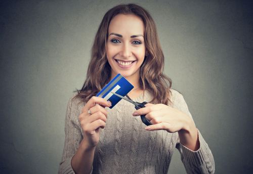 young woman with long brown hair wearing beige sweater smiling while cutting up her credit card in front of her