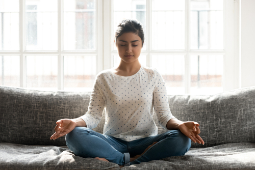young woman sat meditating on grey couch in front of window